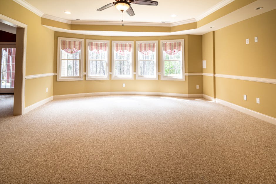 A spacious, empty living room with beige walls and a cream-colored carpet flooring. The room features a large bay window with five tall, white-framed glass panes, decorated with red-and-white striped valances. Natural light illuminates the room, highlighting the clean and well-maintained surfaces. A ceiling fan with a light fixture is centrally mounted on the ceiling, which has recessed lighting. The walls are adorned with white crown molding and a matching chair rail that runs horizontally around the room. The floor appears freshly vacuumed or steam cleaned, emphasizing the surface cleaning and deep cleaning services offered by Harrow Carpet Cleaning. The room is free of dust or clutter, demonstrating good hygiene and maintenance, ideal for domestic cleaning or sanitisation purposes. Visible electrical outlets and a doorway leading to another room add to the overall neat appearance of this residential space.