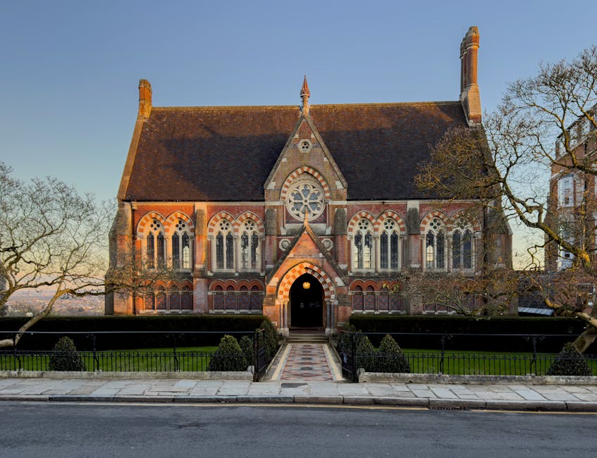 A large historic stone church with intricate Gothic architecture, featuring tall arched windows, a prominent rose window, and a steeply pitched roof topped with two chimneys. The church is surrounded